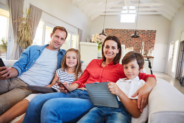 Αυτή η οικογένεια λατρεύει τις συσκευές της. Πορτρέτο μιας χαμογελαστής οικογένειας που κάθεται μαζί στον καναπέ του σαλονιού τους χρησιμοποιώντας διάφορα μέσα και συσκευές - Φωτογραφία, εικόνα