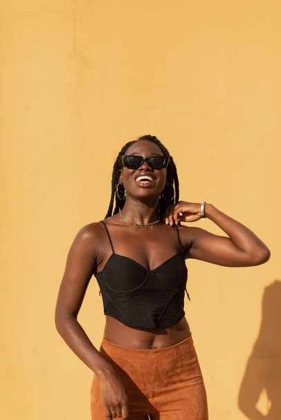 Portrait of a young black female posing with style and smiling with a plain orange wall behind her during a summer sunset - Photo, Image