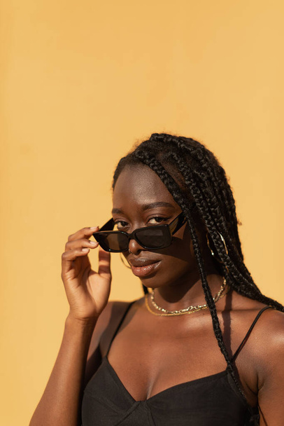 Close-up portrait of a young black woman looking at camera with expressive gaze over glasses during sunset - Fotoğraf, Görsel