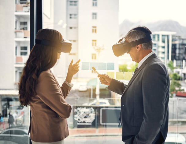There are many ways to perceive success. two businesspeople wearing VR headsets while working with notes on a glass wall in an office - Foto, afbeelding
