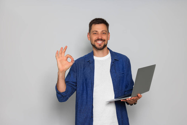 Smiling man with laptop showing okay gesture on light grey background - Photo, Image