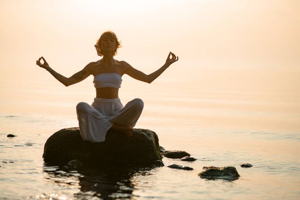 Silhouette of beautiful woman practicing yoga in lotus pose sitting on stone. Healthy life concept. - Фото, изображение