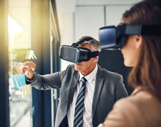 Reaching out to where it was thought impossible before. two businesspeople wearing VR headsets while working with notes on a glass wall in an office - Foto, Bild