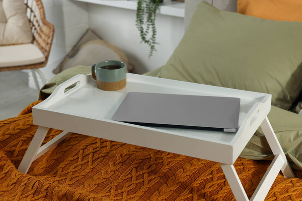 Modern dizüstü bilgisayarlı beyaz tepsi ve yatağın içinde bir fincan aromatik çay. - Fotoğraf, Görsel