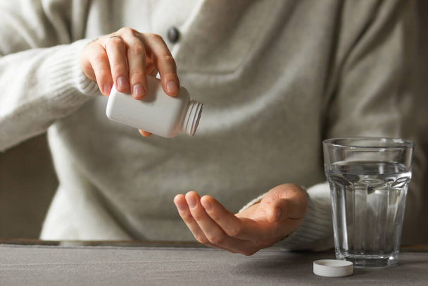 Крупным планом человек руки держит белую пластиковую бутылку с таблетками и готов принять дополнение таблетки со стаканом воды. Больной человек принимает лекарства. Больной человек нуждается в лекарстве от головной боли и простуды. - Фото, изображение