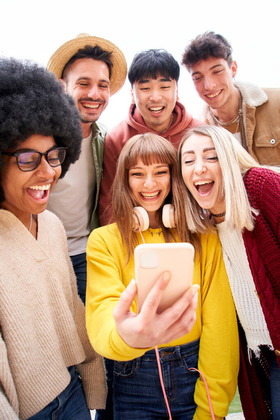 Vertical photo. Multicultural group of friends using cell phones and laughing. Cheerful students lined up and looking at smartphone screen Young people smiling and looking at their mobile High quality - Photo, image