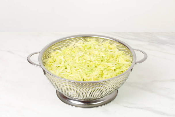 Grated salted zucchini placed in a steel microperforated colander, for straining out the water of them, over marble white kitchen board. Preparing marrow vegetables for freezing or for cooking. Copy space - Photo, image