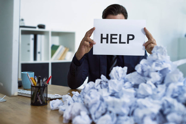 Assistance please. a stressed out businessman sitting at his desk overwhelmed by paperwork holding up a help sign - Photo, image