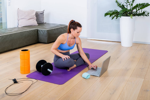 Fit young woman in sports clothes searching for training video course at web using laptop, lying on floor yoga mat in the modern living room with gym equipment - Foto, Imagem