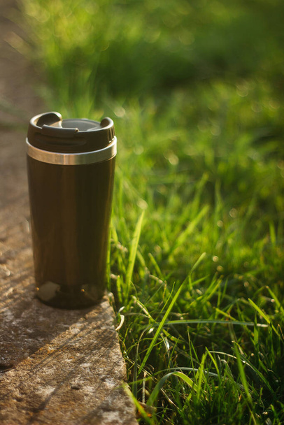 a thermos for hot drinks on a hike or a walk in the park. a thermos on the background of the sunset in the garden. - Photo, Image