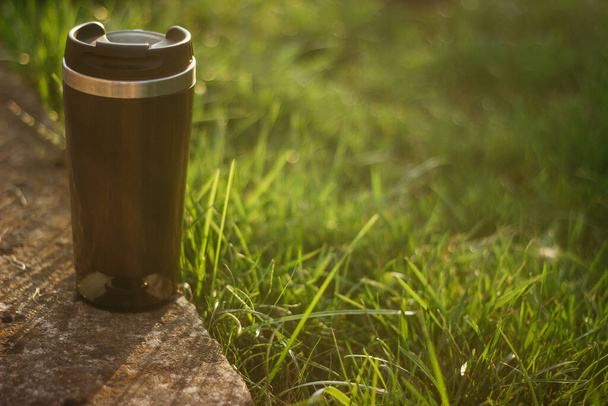 a thermos for hot drinks on a hike or a walk in the park. a thermos on the background of the sunset in the garden. - Valokuva, kuva