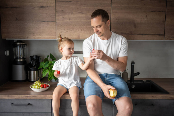 Cute little girl and her handsome dad are eating fruit in modern kitchen. Healthy eating. High quality photo - Photo, Image