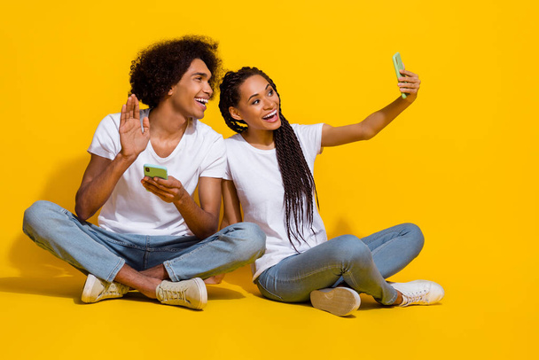 Full size photo of two cheerful people sit floor make selfie arm waving hi isolated on yellow color background. - Φωτογραφία, εικόνα