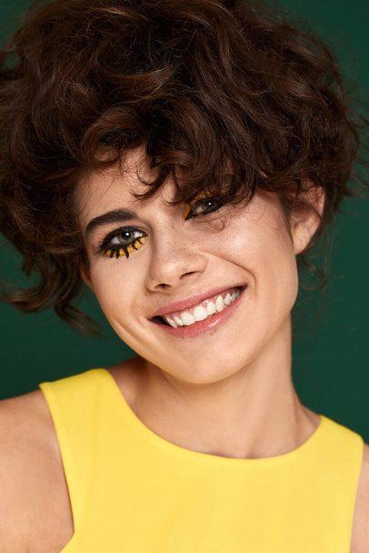 young curly woman with creative make-up in yellow dress on green background. - Foto, afbeelding