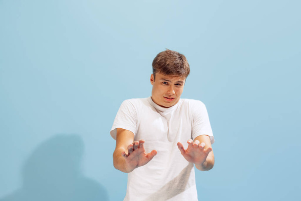 Portrait of young man in white T-shirt posing, showing unpleasant emotions isolated over blue studio background. Escaping, avoiding. Concept of emotions, facial expression, lifestyle, fashion, youth - Photo, Image