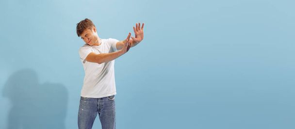 Portrait of young man in casual T-shirt and jeans posing isolated over blue background. Stretching out hands to avoid something. Concept of emotions, facial expression, lifestyle, fashion, youth - Fotoğraf, Görsel