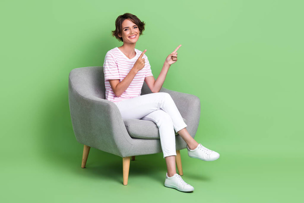 Full length photo of pretty young girl sit chair fingers point empty space wear stylish striped garment isolated on green color background. - Foto, imagen