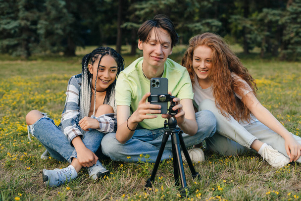 Группа молодых влиятельных подростков, сидящих на траве в парке и снимающих живое видео в Интернете. Концепция дружбы и новых технологий в летнее время - Фото, изображение
