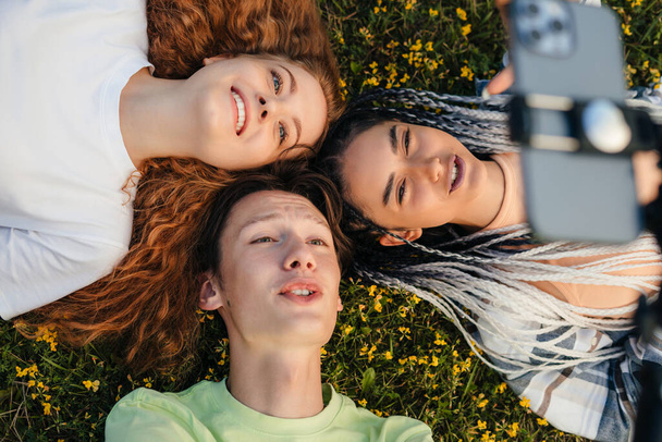 Top view of two young girls and a young boy lying on the green grass in the park, holding their heads together trying to take an interesting picture to post on - Foto, Imagem
