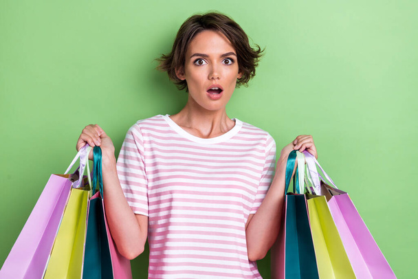 Photo of young adorable funny face open mouth girl wear pink striped t-shirt hold shopping bags shocked prices black friday isolated on green color background. - Foto, Imagem