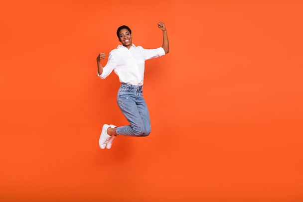 Полноразмерное фото радостной позитивной женщины-прыгуньи поднимает кулаки в успехе триумф изолирован на оранжевом фоне цвета. - Фото, изображение