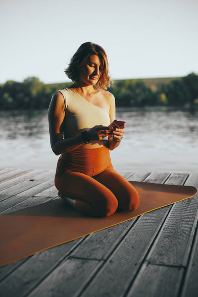A fit woman is kneeling on a yoga mat and texting messages on the phone on the dock. - Photo, Image