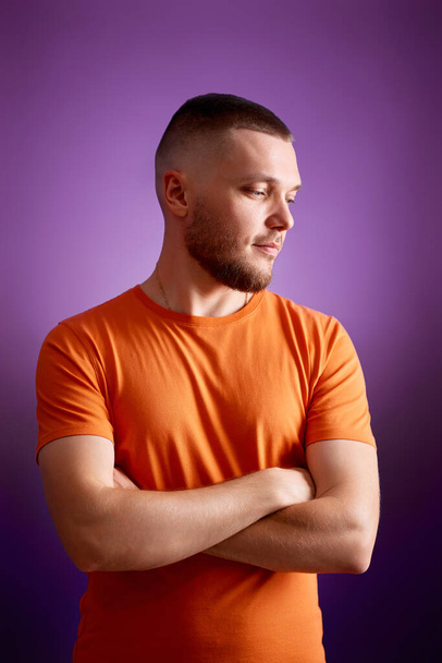 young man in orange t-shirt posing with crossed arms on purple studio background - Foto, Imagem
