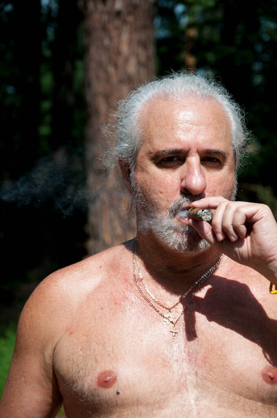 пожилой мужчина курит кубинскую сигару наслаждаясь летними каникулами и загаром на пенсии. - Фото, изображение