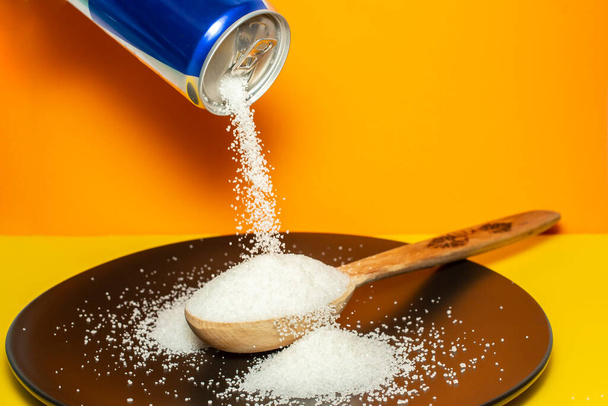 Pouring crystal sugar from a blue can to a wooden spoon on a black plate and orange background.  Motion blur on sugar texture. Sugar danger concept - Foto, Imagen