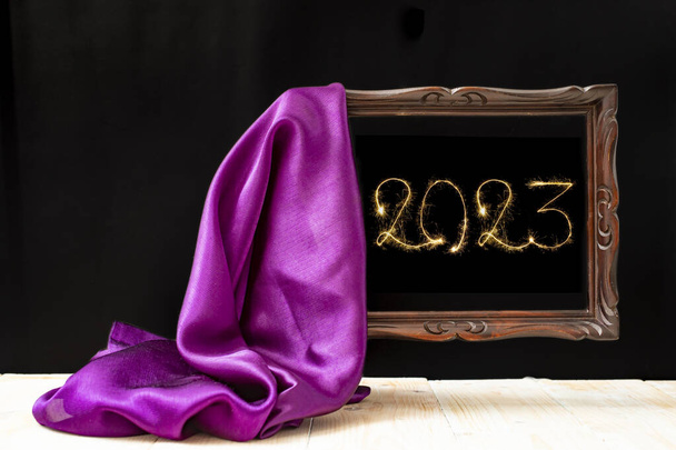 2023 число, написанное с искрой внутри коричневой рамки. Фиолетовая атласная ткань обнажает винтажную деревянную рамку на черном фоне. С наступающим Новым годом!. - Фото, изображение
