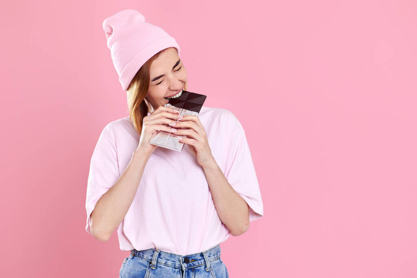 charming woman in casual t-shirt biting sweet chocolate bar isolated on pastel pink background - Photo, image