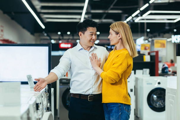 Happy diverse young family man and woman choose electronic household appliances, shop for new apartment in supermarket - Photo, Image