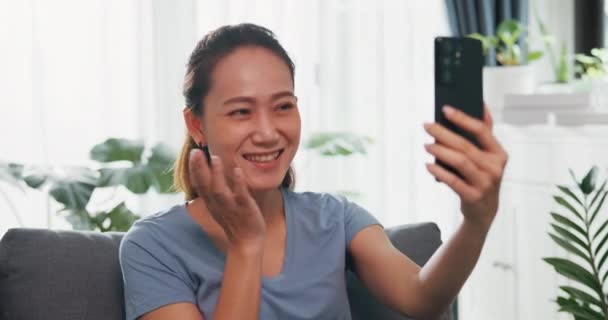Close-up Young Asian woman wear casual site on couch use smartphone raise hand video call to say hello and talk to family and friend with happiness and smiling in living room indoor plants at home. - Footage, Video