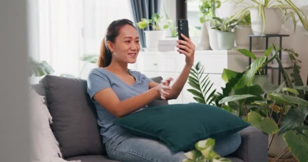 Young Asian woman wear casual site on couch use smartphone raise hand video call to say hello and talk to family and friend with happiness and smiling in living room indoor plants at home. - Footage, Video