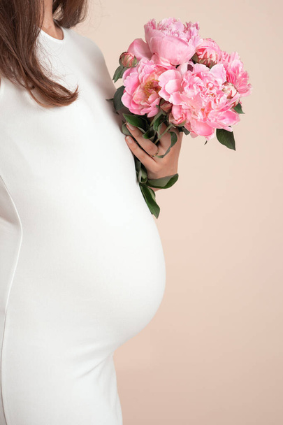 A body portrait of a young white pregnant female in white dress holding pink peonies on beige colored background - 写真・画像