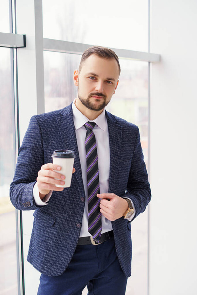 A young man in a business suit with a cup of coffee stands at a large window and looks out. Young businessman on the break - Photo, Image