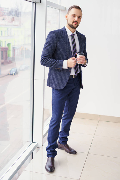 Молодой человек в деловом костюме с чашкой кофе стоит у большого окна и смотрит в окно. Молодой бизнесмен на перерыве - Фото, изображение