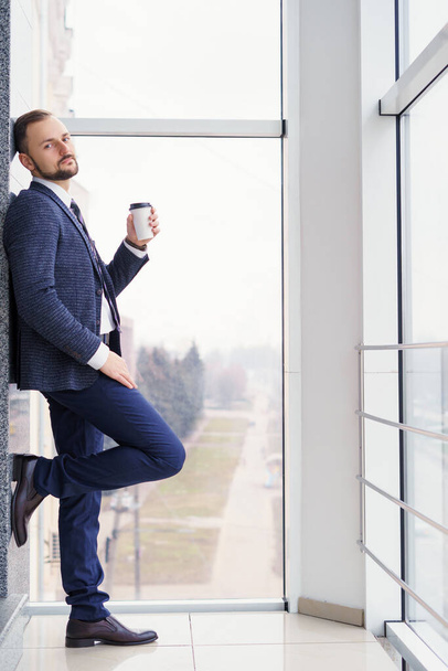Ένας νεαρός με ένα κοστούμι με ένα φλιτζάνι καφέ στέκεται σε ένα μεγάλο παράθυρο και κοιτάζει έξω. Νεαρός επιχειρηματίας στο διάλειμμα - Φωτογραφία, εικόνα