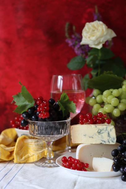 Τυρί καμαμπέρ, πράσινα και μαύρα σταφύλια, ένα ποτήρι ροζέ κρασί σε ένα τραπέζι. Πολύχρωμο καλοκαίρι νεκρή φύση με εποχιακά φρούτα και μούρα.  - Φωτογραφία, εικόνα