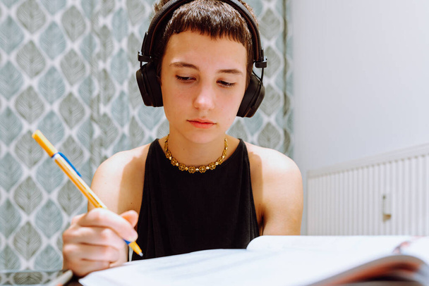 Concentrated millennial teenager girl in headphones sits at table at home, takes notes in notebook, learning online, focused young woman, pensive look, distance education concept. Homeschooling, homework, university student - Foto, immagini