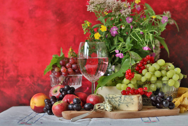 Ένα ποτήρι ροζ κρασί, σταφύλια, μούρα και λουλούδια σε ένα τραπέζι. Πεντανόστιμο καλοκαιρινό φαγητό από κοντά. Υφές φόντου με χώρο αντιγραφής. - Φωτογραφία, εικόνα