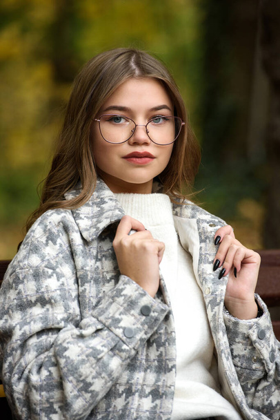 Close-up portrait of young woman in a stylish trench coat and in a white sweater resting on the bench. warm autumn day. warm autumn day. brown wooden bench - Foto, Imagen
