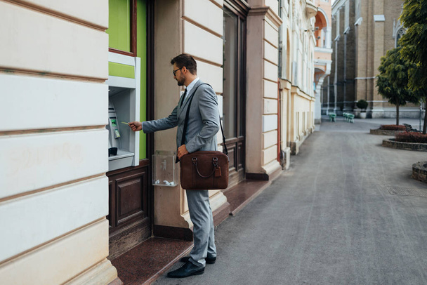 Handsome middle age businessman with eyeglasses standing on city street and using ATM machine to withdraw money from credit or debit card.  - Foto, imagen