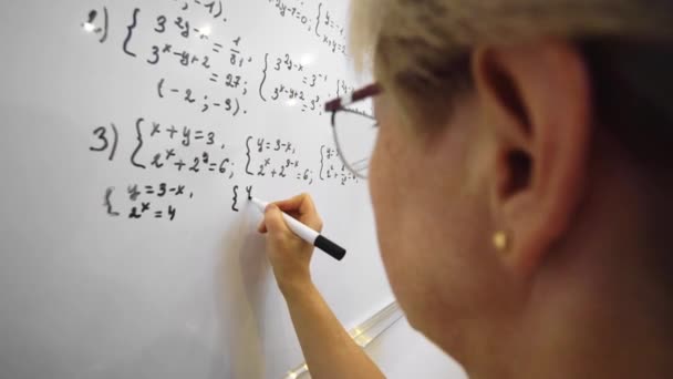 Math algebra lesson, education. Aged teacher in glasses is solving maths equation on whiteboard close up. - Video