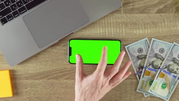 The man is zooming in the phone green screen mockup in horizontal position. 100 dollar banknotes on the table. - Video, Çekim