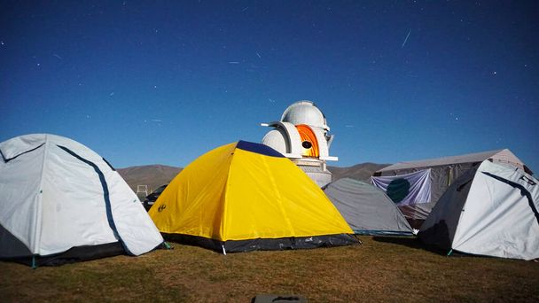 A camp with colored tents near the observatory. The Perseid starfall. A large teleskom watches the starry night sky. Meteorites and planes fly by, leaving traces in the sky. The stars are burning. - Foto, afbeelding