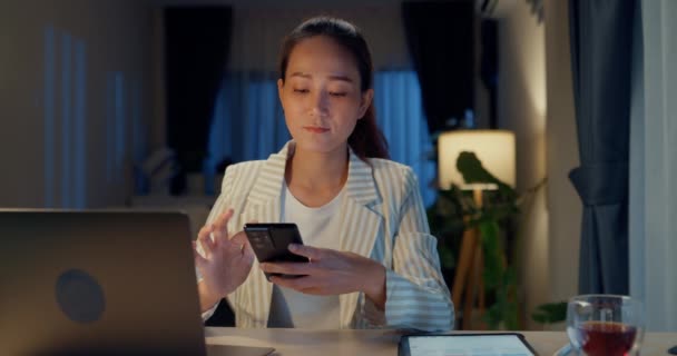 若い幸せなアジアのビジネスマンの女性は、自宅の夜に居心地の良いリビングルームで休息を取るスマートフォンにノートパソコンのコンピュータ入力メッセージと机の前に正式な座席を着用してください。残業の概念. - 映像、動画