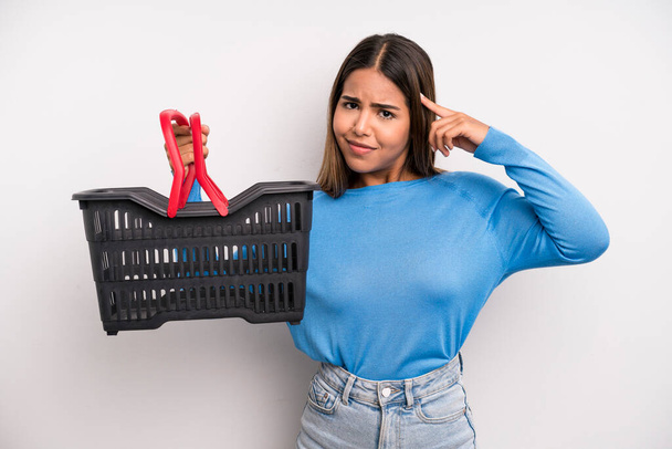hispanic pretty woman feeling confused and puzzled, showing you are insane. empty supermarket basket concept - Фото, изображение