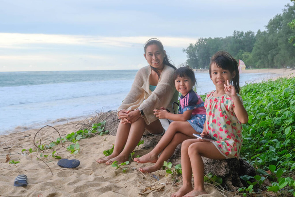 Ευτυχισμένη ασιατική οικογένεια της μητέρας και των κορών κάθεται σε ένα κούτσουρο χαμογελώντας στην κάμερα στην παραλία κατά τη διάρκεια των καλοκαιρινών διακοπών στο ηλιοβασίλεμα. Οικογενειακό καλοκαιρινό ταξίδι στην παραλία. έννοια ταξίδια και διακοπές. - Φωτογραφία, εικόνα