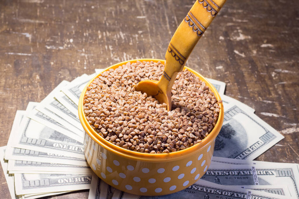 Buckwheat in a bowl with a wooden spoon on dollar bills. Rising prices in Ukraine due to the war - Foto, Bild